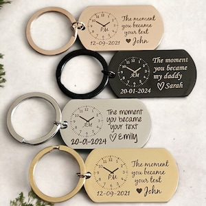 May include: Four silver, black, and gold keychains with a clock face and engraved text. The text reads "The moment you became your text" with a heart and name. The clock face shows the time and AM or PM. The date is also engraved on each keychain.
