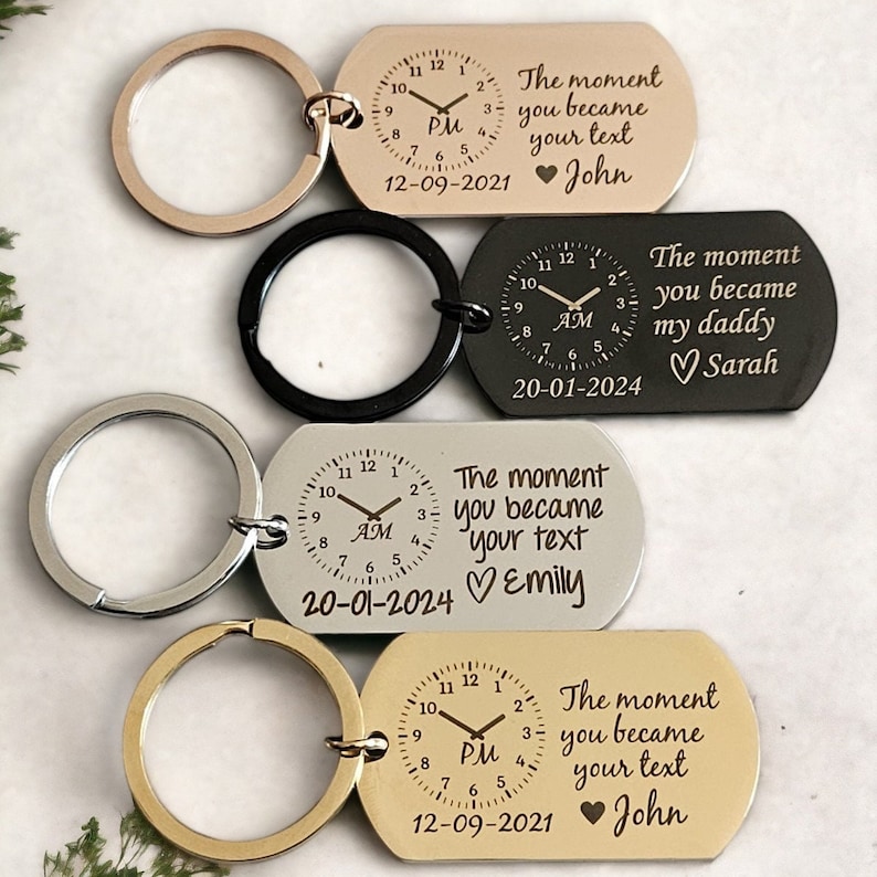 May include: Four silver, black, and gold keychains with a clock face and engraved text. The text reads "The moment you became your text" with a heart and name. The clock face shows the time and AM or PM. The date is also engraved on each keychain.