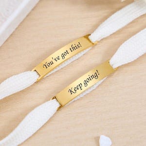 May include: Two white shoelaces with gold-colored rectangular tags. One tag reads "You've got this!" and the other reads "Keep going!" The laces are on a light wooden surface.