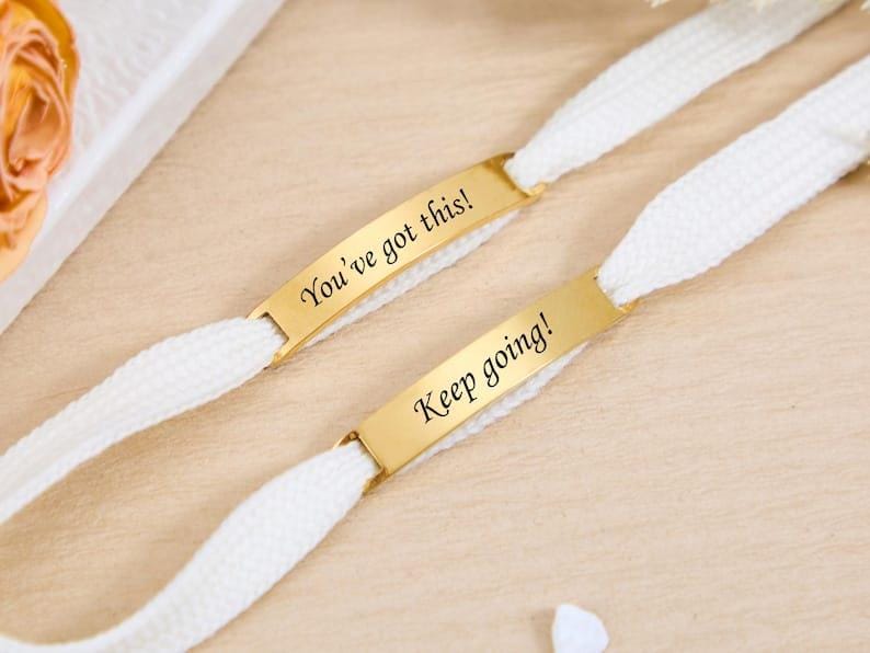May include: Two white shoelaces with gold-colored rectangular tags. One tag reads "You've got this!" and the other reads "Keep going!" The laces are on a light wooden surface.