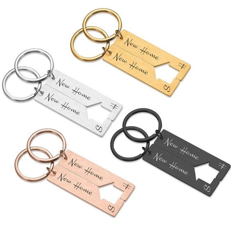 May include: Two sets of matching keychains with interlocking key rings. Each keychain has a silver, gold, or black metal tag with the words "New Home" engraved on it. The tags are shaped like a house with a keyhole.