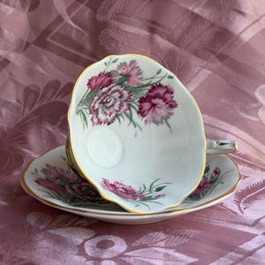 May include: A white porcelain teacup and saucer set featuring a delicate floral pattern of magenta and pink carnations with green foliage. The edges of both the cup and the saucer are finished with a thin, elegant gold trim along the scalloped rims.