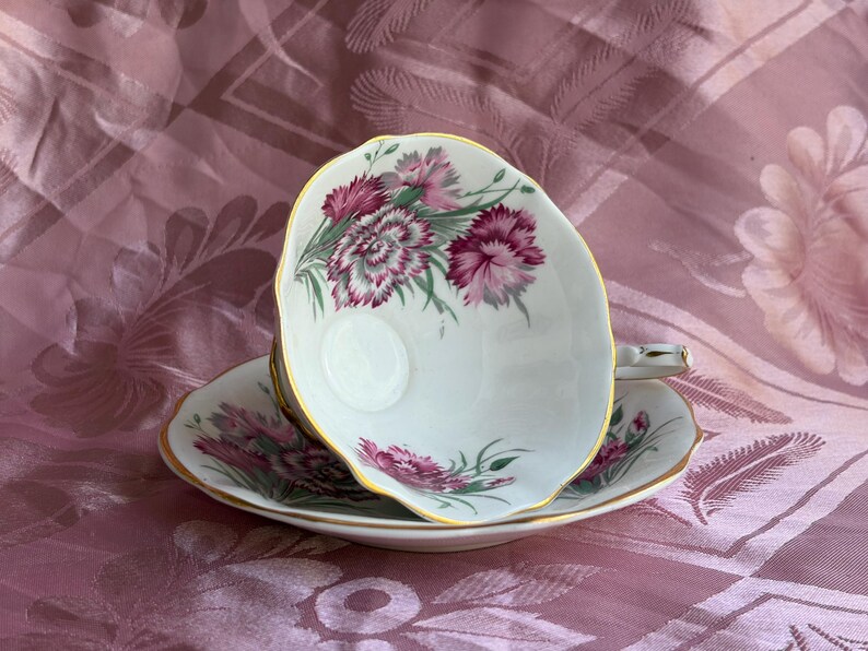 May include: A white porcelain teacup and saucer set featuring a delicate floral pattern of magenta and pink carnations with green foliage. The edges of both the cup and the saucer are finished with a thin, elegant gold trim along the scalloped rims.