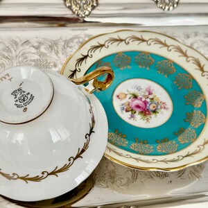 May include: A vintage teacup and saucer set. The white teacup has gold trim and a gold handle. The saucer is turquoise with gold accents and a floral center. The set is likely made of porcelain.