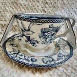 May include: A ceramic teacup and saucer set featuring a dark blue floral and bird pattern on a cream background. The rim of both the cup and saucer is decorated with a dark blue geometric border. The set is held together by a clear plastic display frame.