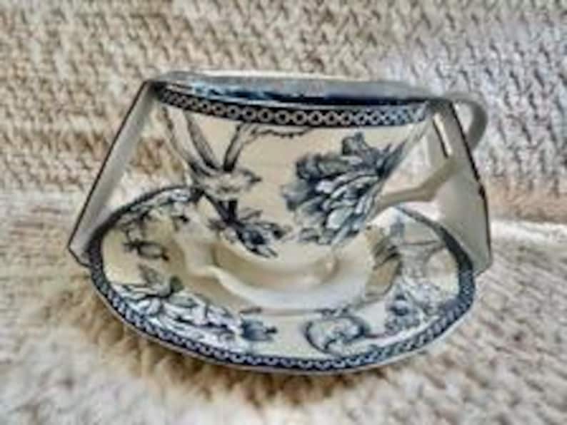 May include: A ceramic teacup and saucer set featuring a dark blue floral and bird pattern on a cream background. The rim of both the cup and saucer is decorated with a dark blue geometric border. The set is held together by a clear plastic display frame.