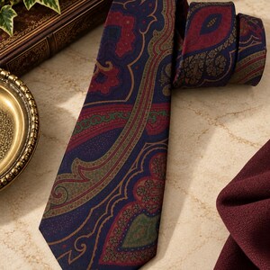 Pode incluir: Uma gravata com base azul-escura e um padr&atilde;o paisley intrincado em tons de vermelho profundo, verde-azeitona e dourado. O tecido apresenta uma textura lisa com uma ponta afiada e uma sec&ccedil;&atilde;o superior dobrada com o mesmo design.