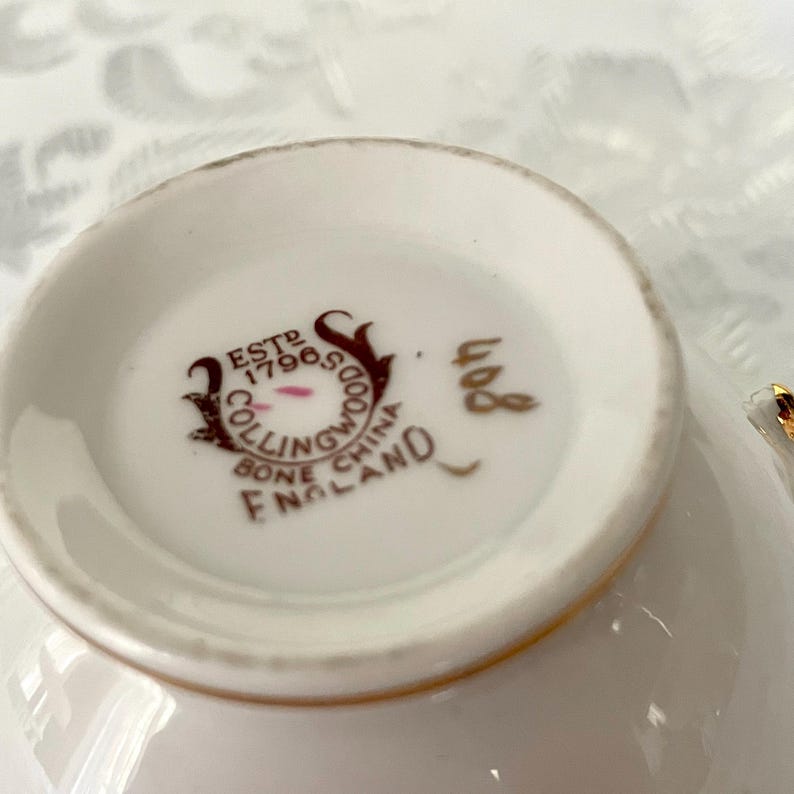 Puede incluir: Primer plano del interior de una taza de t&eacute; blanca con borde dorado. La base de la taza presenta un logotipo marr&oacute;n y dorado que dice "ESTD 17965 COLLINGWOOD BONE CHINA ENGLAND". El n&uacute;mero "408" est&aacute; impreso en dorado.