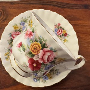 May include: A white teacup and saucer with a floral pattern in shades of pink, yellow, and green. The floral design features roses, daisies, and other flowers.