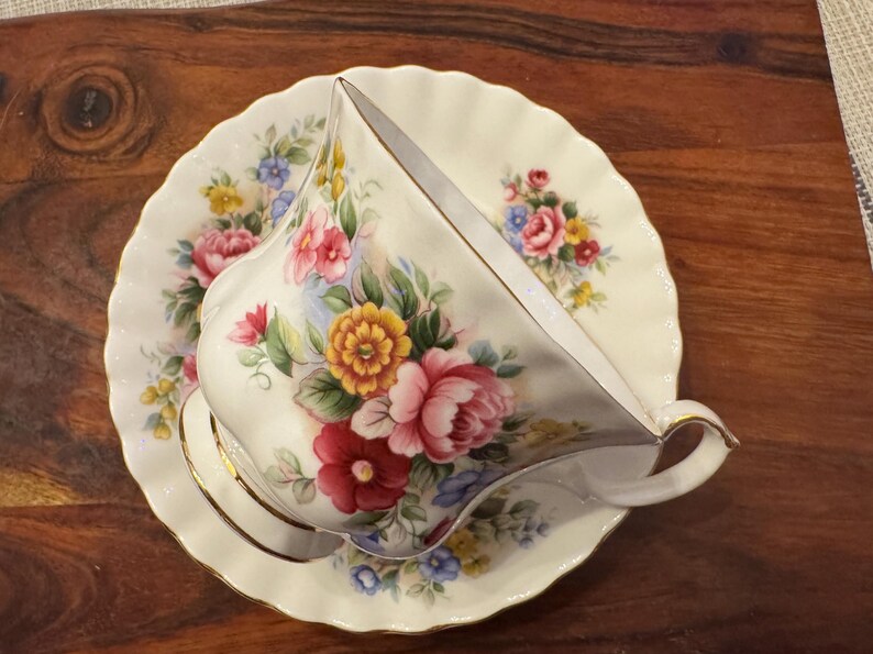 May include: A white teacup and saucer with a floral pattern in shades of pink, yellow, and green. The floral design features roses, daisies, and other flowers.