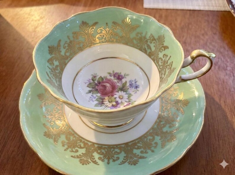 Peut inclure: Un ensemble tasse et soucoupe en porcelaine avec une bordure vert menthe p&acirc;le orn&eacute;e de filigranes dor&eacute;s complexes. Le centre de la tasse pr&eacute;sente un bouquet floral de roses roses, de violettes violettes et de marguerites blanches.