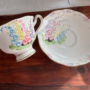 May include: A white teacup and saucer set with a floral design. The cup and saucer feature a colorful floral pattern with pink, blue, yellow, and green accents. The floral design is reminiscent of a bouquet of flowers.