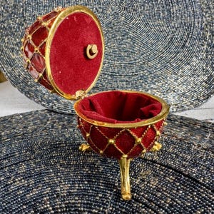 May include: A red and gold jeweled egg-shaped trinket box with a hinged lid and three gold feet. The box is open and lined with red velvet.