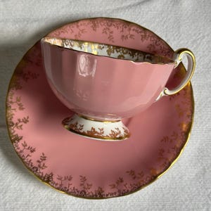 Peut inclure: Une tasse et une soucoupe roses avec des bordures dor&eacute;es et des motifs floraux. La tasse a un design cannel&eacute; et repose sur une soucoupe assortie. L'ensemble est probablement vintage.