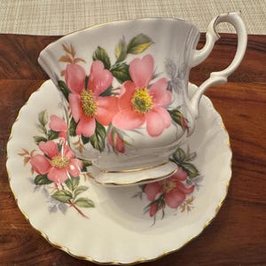 Puede incluir: Un juego de taza y platillo de t&eacute; blanco con un dise&ntilde;o floral con rosas rosas. La taza de t&eacute; tiene un asa delicada y un borde dorado alrededor del borde.
