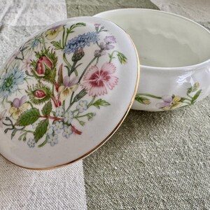 Op de afbeelding: Witte keramische sieradendoos met een bloemmotief op het deksel. Het deksel heeft een boeket van kleurrijke bloemen, waaronder roze, blauwe en gele bloemen. De doos heeft een gouden rand en een bijpassend bloemmotief op de basis.