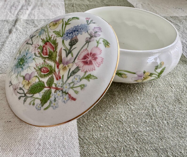 Op de afbeelding: Witte keramische sieradendoos met een bloemmotief op het deksel. Het deksel heeft een boeket van kleurrijke bloemen, waaronder roze, blauwe en gele bloemen. De doos heeft een gouden rand en een bijpassend bloemmotief op de basis.