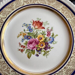 May include: A white ceramic plate with a floral bouquet design. The bouquet features a red tulip, pink rose, and various other colorful flowers. The plate has a gold rim and a blue border with decorative patterns.