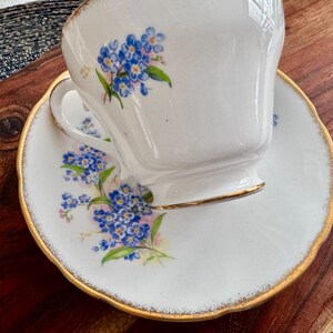 May include: A white teacup and saucer set with a delicate floral pattern of blue forget-me-nots. The cup and saucer have a gold trim around the edges.