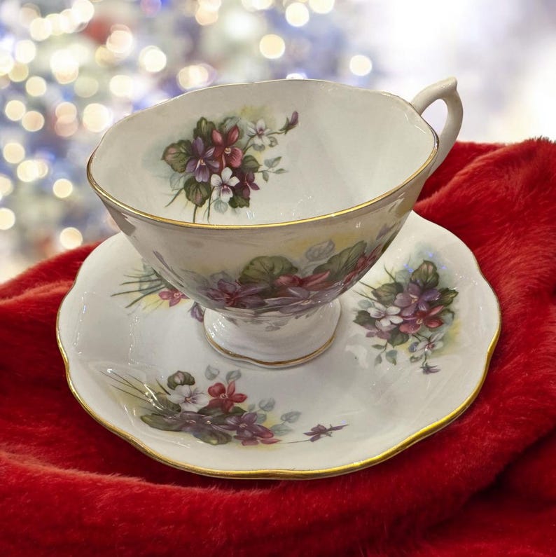 May include: A white teacup and saucer with a floral design featuring purple and white flowers. The cup and saucer have a gold trim.