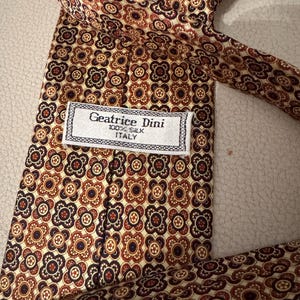 May include: A necktie featuring a repeating geometric floral pattern in shades of dark red, cream, and navy blue. A white fabric label is stitched to the back, displaying the text Geatrice Dini, 100% SILK, and ITALY in black lettering.