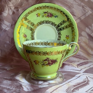 May include: A light green porcelain teacup and matching saucer set featuring a gold leaf garland pattern and a central floral bouquet of pink, yellow, and purple flowers. The teacup has a curved handle and gold trim along the rim and base.