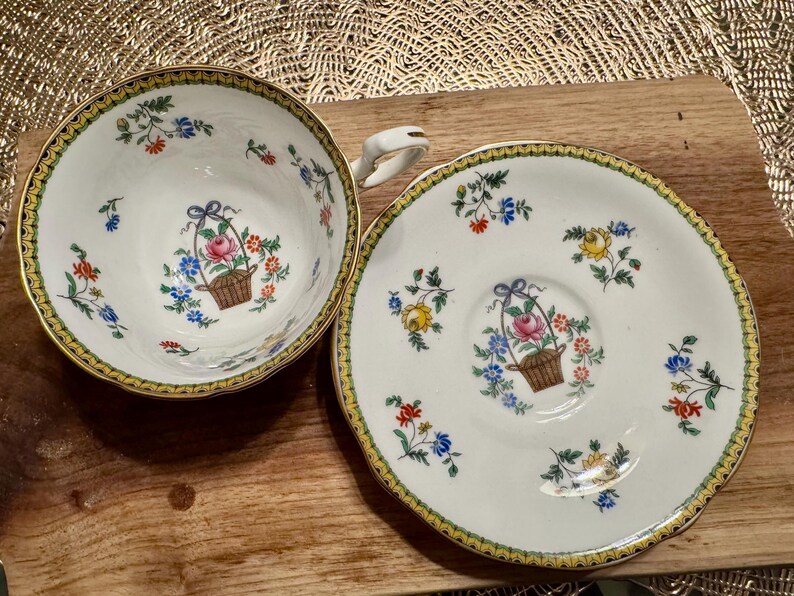 Pu&ograve; includere: Un set da t&egrave; vintage composto da una tazza e un piattino con un motivo floreale. La tazza e il piattino sono bianchi con un bordo dorato e presentano un delicato disegno floreale con fiori rosa, gialli e blu. Il disegno include un cestino di fiori con un fiocco.