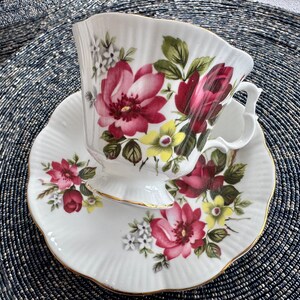 Puede incluir: Un juego de taza y platillo de t&eacute; blanco con un patr&oacute;n floral con flores rosas, rojas, amarillas y verdes. El juego tiene un borde dorado.