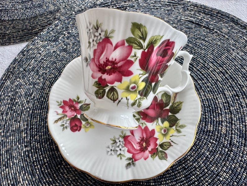 Puede incluir: Un juego de taza y platillo de t&eacute; blanco con un patr&oacute;n floral con flores rosas, rojas, amarillas y verdes. El juego tiene un borde dorado.