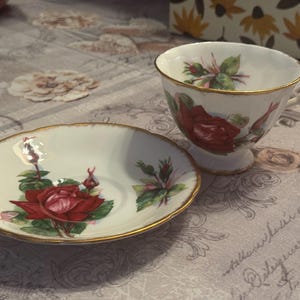 May include: A white teacup and saucer set with a red rose floral design and gold trim. The teacup has a handle and sits next to the matching saucer. The set is on a patterned surface.