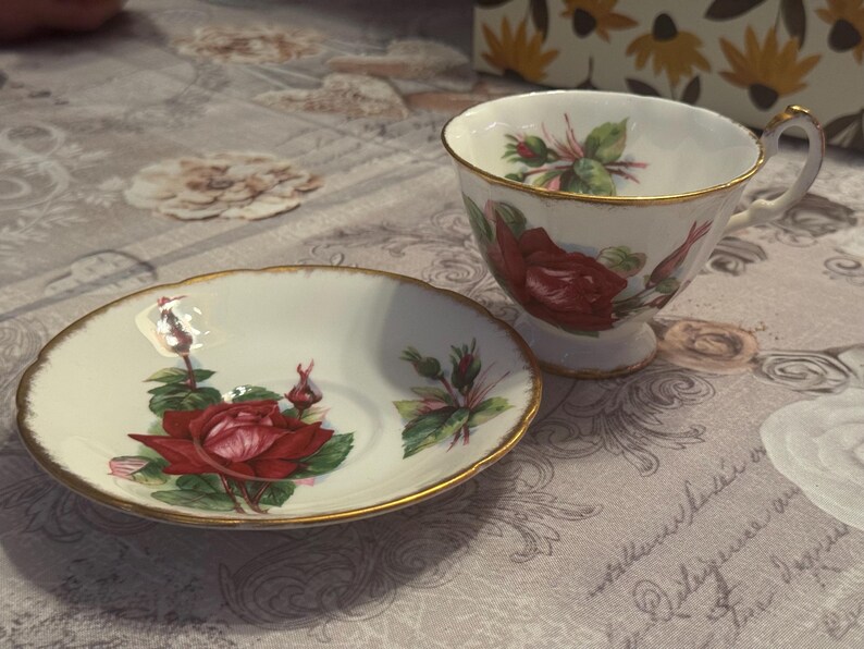May include: A white teacup and saucer set with a red rose floral design and gold trim. The teacup has a handle and sits next to the matching saucer. The set is on a patterned surface.