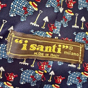 May include: A dark blue necktie featuring a repeating pattern of blue and white checkered pigs wearing yellow hats and red bows, red watering cans, and white garden rakes. A brown fabric label reads i santi milano MADE IN ITALY.