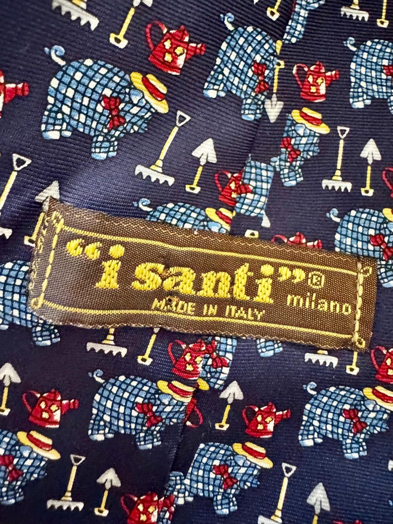 May include: A dark blue necktie featuring a repeating pattern of blue and white checkered pigs wearing yellow hats and red bows, red watering cans, and white garden rakes. A brown fabric label reads i santi milano MADE IN ITALY.