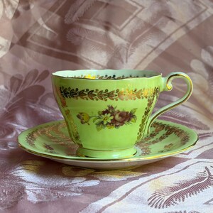 May include: A light green porcelain teacup and saucer set featuring a scalloped rim, a gold-toned handle, and intricate gold leaf detailing along the edges. The center of the cup displays a small, colorful floral bouquet with red and yellow petals.