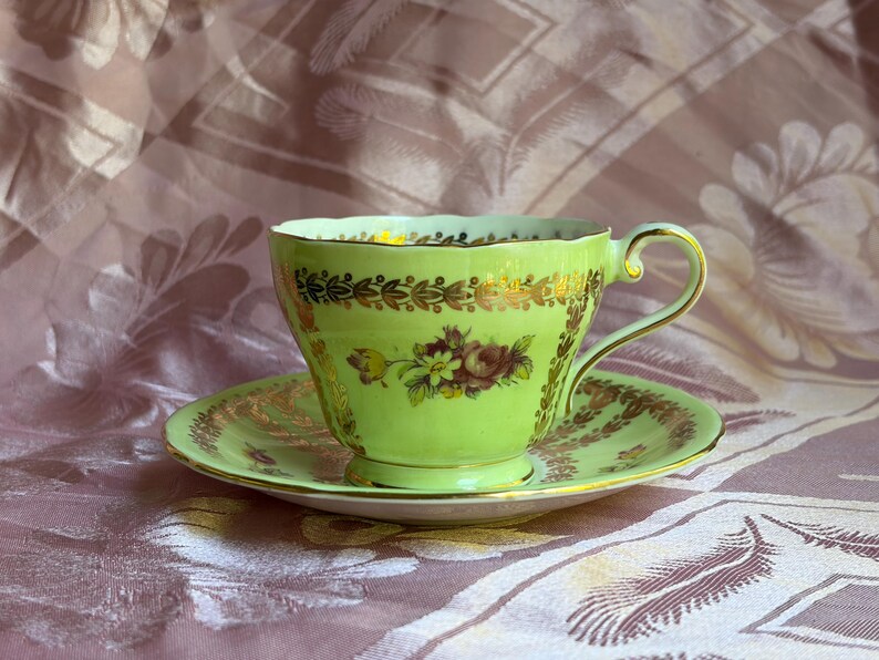 May include: A light green porcelain teacup and saucer set featuring a scalloped rim, a gold-toned handle, and intricate gold leaf detailing along the edges. The center of the cup displays a small, colorful floral bouquet with red and yellow petals.