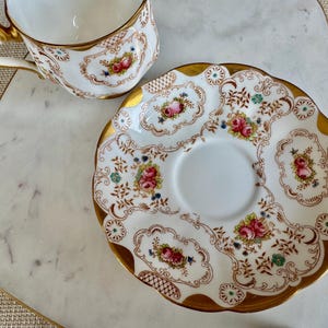 K&ouml;nnte beinhalten: Ein wei&szlig;es Tee-Service mit Tasse und Untertasse. Der Rand und der Griff sind goldfarben. Das florale Design zeigt rosa Rosen, blaue Akzente und braune Details. Die Untertasse hat einen gewellten Rand.