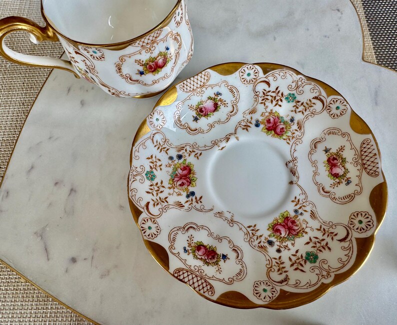 K&ouml;nnte beinhalten: Ein wei&szlig;es Tee-Service mit Tasse und Untertasse. Der Rand und der Griff sind goldfarben. Das florale Design zeigt rosa Rosen, blaue Akzente und braune Details. Die Untertasse hat einen gewellten Rand.