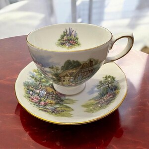 May include: A white teacup and saucer set with a gold rim. The teacup and saucer feature a painted scene of a cottage in a green field with flowers and trees.