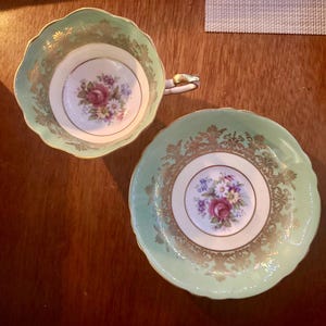 Peut inclure: Une tasse &agrave; th&eacute; en porcelaine et sa soucoupe assortie, dot&eacute;es d'une bordure vert menthe p&acirc;le avec des d&eacute;tails en filigrane dor&eacute;. Le centre des deux pi&egrave;ces pr&eacute;sente un bouquet floral avec des roses roses et de petites fleurs blanches et bleues.