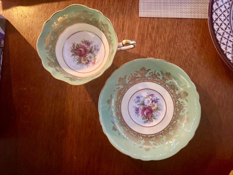 Peut inclure: Une tasse &agrave; th&eacute; en porcelaine et sa soucoupe assortie, dot&eacute;es d'une bordure vert menthe p&acirc;le avec des d&eacute;tails en filigrane dor&eacute;. Le centre des deux pi&egrave;ces pr&eacute;sente un bouquet floral avec des roses roses et de petites fleurs blanches et bleues.
