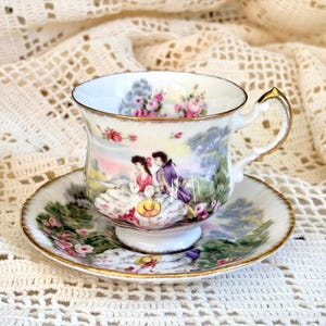 May include: A white porcelain teacup and saucer set featuring a colorful scene of two figures in period clothing amidst a landscape with pink roses. The rim of the cup and the saucer are accented with a thin gold trim and a decorative handle.