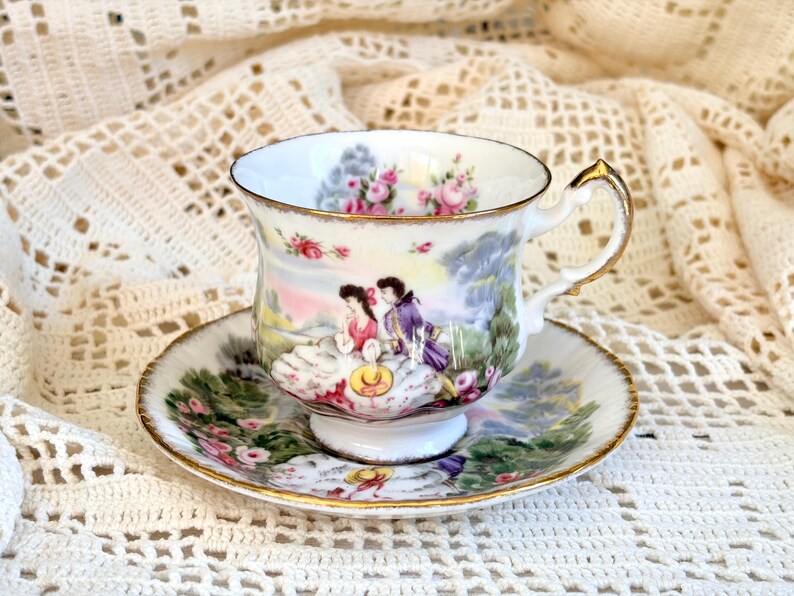 May include: A white porcelain teacup and saucer set featuring a colorful scene of two figures in period clothing amidst a landscape with pink roses. The rim of the cup and the saucer are accented with a thin gold trim and a decorative handle.