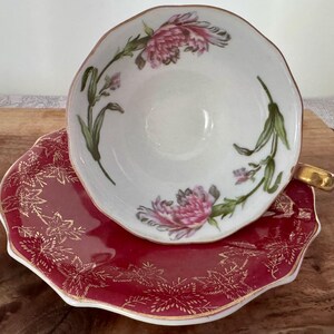 May include: A white teacup with pink floral designs and a gold rim sits atop a red saucer with gold leaf patterns. The teacup has a gold handle. The set is likely vintage and suitable for tea parties.