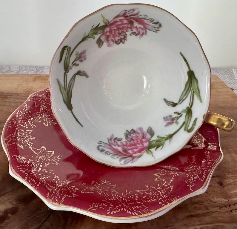 May include: A white teacup with pink floral designs and a gold rim sits atop a red saucer with gold leaf patterns. The teacup has a gold handle. The set is likely vintage and suitable for tea parties.