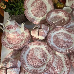 Pu&ograve; includere: Una collezione di piatti antichi in ceramica bianca e rossa, tra cui piatti, ciotole, tazze e una teiera. I piatti presentano un design dettagliato di paesaggio rurale. Il set &egrave; disposto su una superficie bianca.