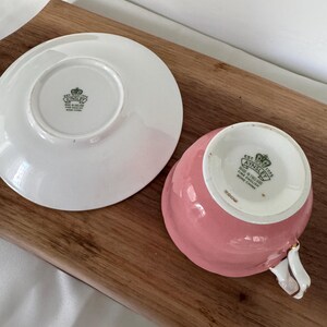 May include: A pink teacup and saucer set, with a white handle and gold trim. The teacup and saucer are made of fine bone china. The saucer is white, and the teacup is a solid pink color. The set is placed on a wooden surface.
