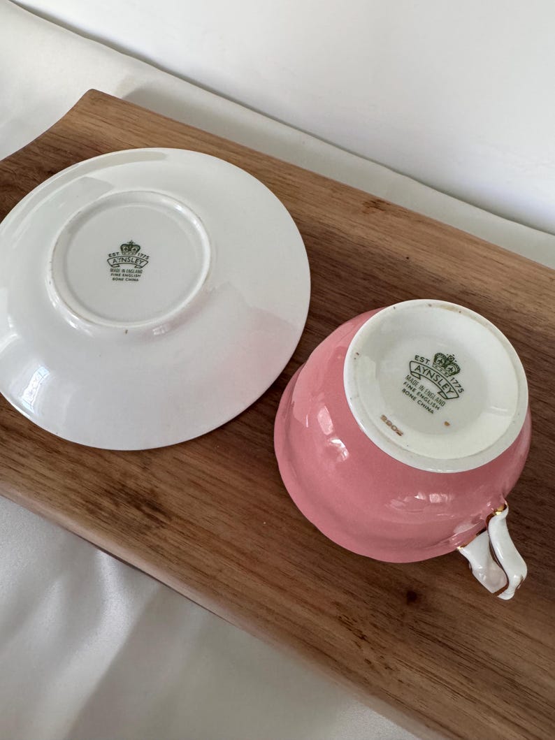 May include: A pink teacup and saucer set, with a white handle and gold trim. The teacup and saucer are made of fine bone china. The saucer is white, and the teacup is a solid pink color. The set is placed on a wooden surface.