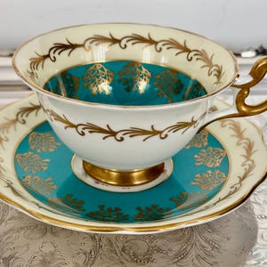 May include: A vintage teacup and saucer set. The teacup and saucer are white with gold trim and teal accents. The interior of the teacup and the center of the saucer are teal with gold floral designs. The handle and base are gold.