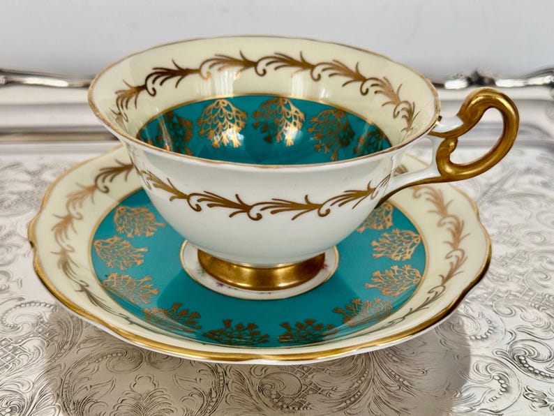 May include: A vintage teacup and saucer set. The teacup and saucer are white with gold trim and teal accents. The interior of the teacup and the center of the saucer are teal with gold floral designs. The handle and base are gold.