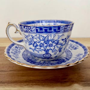May include: A vintage teacup and saucer set. The cup and saucer are white with a blue floral design and gold trim. The teacup has a curved handle. The saucer is round with a scalloped edge.
